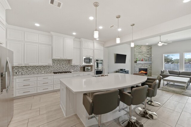 kitchen with a stone fireplace, an island with sink, stainless steel appliances, pendant lighting, and white cabinetry