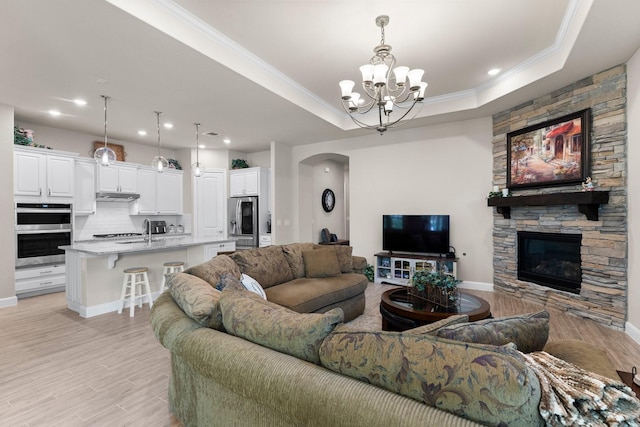 living room with a tray ceiling, a stone fireplace, a chandelier, and light wood-type flooring