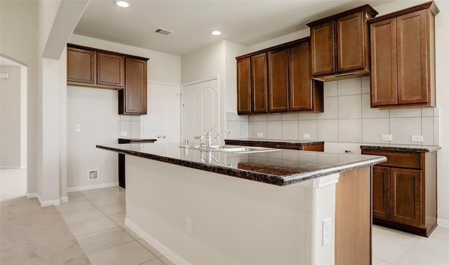 kitchen featuring sink, dark stone countertops, decorative backsplash, light tile patterned floors, and a center island with sink