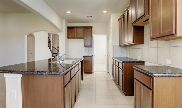 kitchen with sink, an island with sink, decorative backsplash, and dark stone countertops