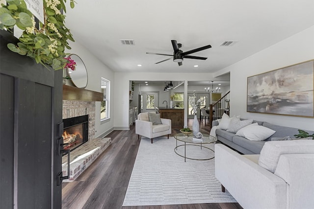 living room with a stone fireplace, dark wood-type flooring, and ceiling fan with notable chandelier