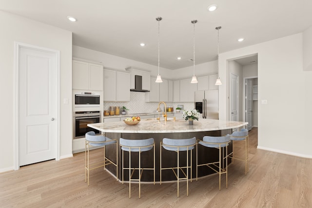 kitchen with pendant lighting, wall chimney exhaust hood, light wood-type flooring, a large island, and stainless steel appliances