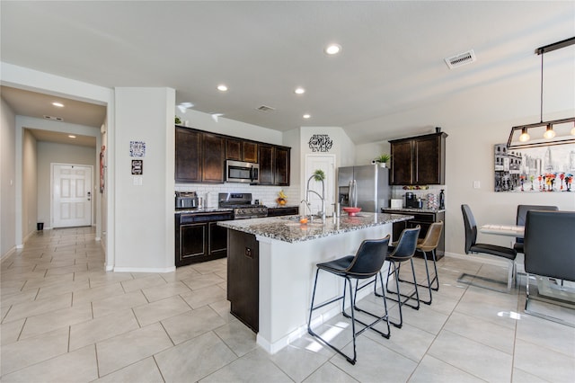 kitchen featuring dark brown cabinets, stainless steel appliances, light stone counters, decorative backsplash, and a kitchen island with sink