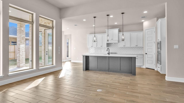 kitchen with white cabinets, light countertops, hanging light fixtures, backsplash, and an island with sink