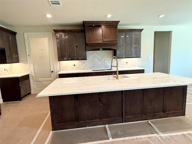 kitchen featuring decorative backsplash, dark brown cabinetry, sink, and an island with sink