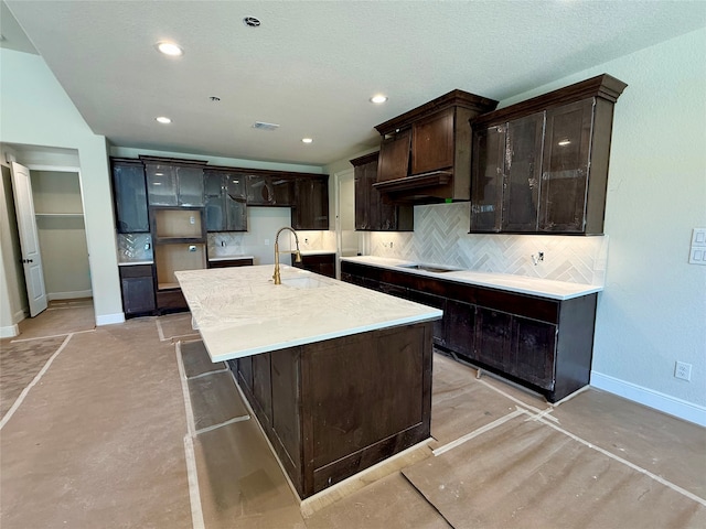 kitchen featuring black electric stovetop, tasteful backsplash, dark brown cabinets, sink, and an island with sink