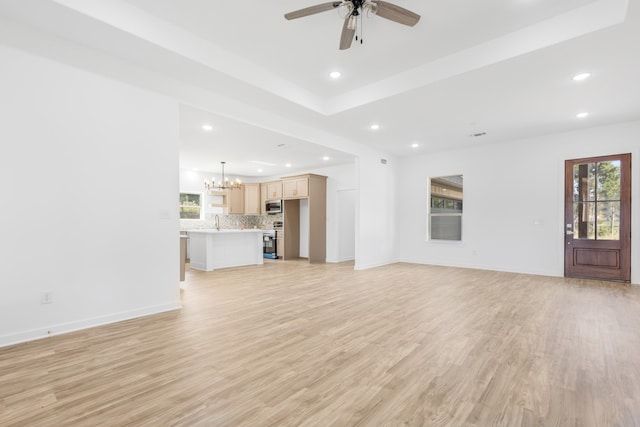 unfurnished living room with light hardwood / wood-style floors, sink, ceiling fan with notable chandelier, and a raised ceiling