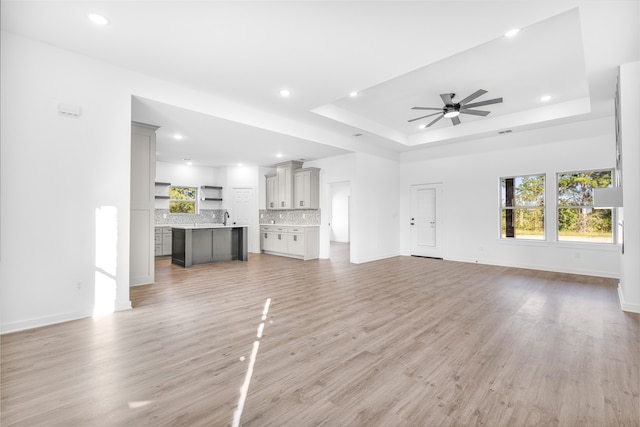 unfurnished living room featuring sink, a tray ceiling, light wood-type flooring, and ceiling fan