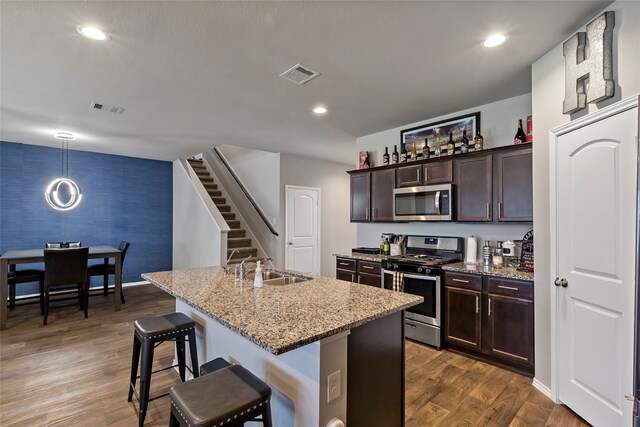 kitchen with a kitchen island with sink, dark wood-type flooring, a kitchen breakfast bar, light stone countertops, and appliances with stainless steel finishes