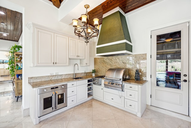 kitchen with sink, white cabinetry, pendant lighting, light stone counters, and premium range hood