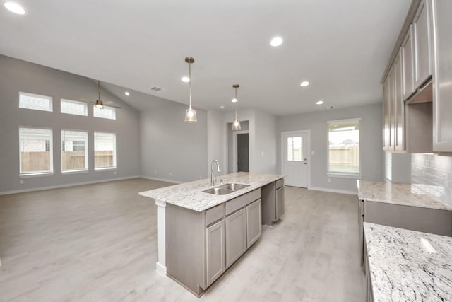 kitchen featuring light wood-type flooring, an island with sink, hanging light fixtures, light stone countertops, and sink