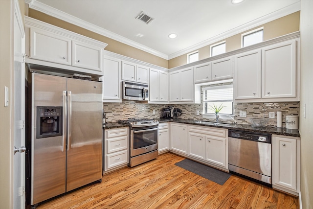 kitchen with dark stone counters, white cabinets, ornamental molding, appliances with stainless steel finishes, and light hardwood / wood-style floors