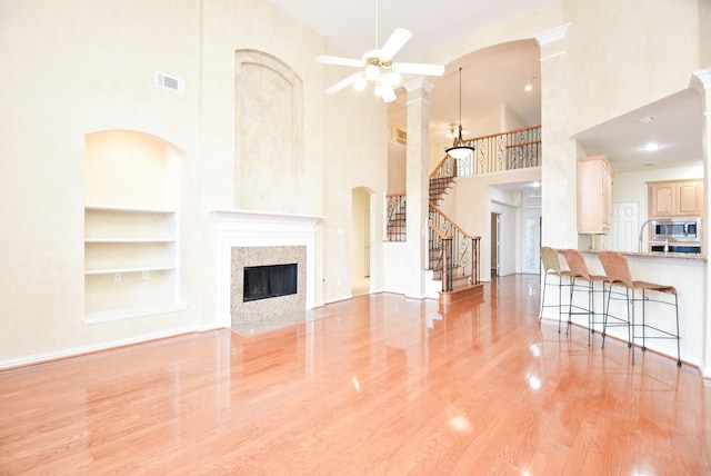 unfurnished living room with a towering ceiling, light hardwood / wood-style floors, ceiling fan, a premium fireplace, and built in shelves