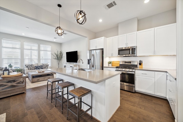 kitchen featuring a kitchen island with sink, stainless steel appliances, hanging light fixtures, and dark wood-type flooring