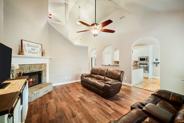 living room with ceiling fan, high vaulted ceiling, a brick fireplace, and light wood-type flooring