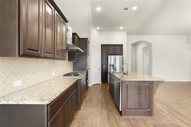 kitchen with an island with sink, light wood-type flooring, wall chimney exhaust hood, dark brown cabinetry, and sink