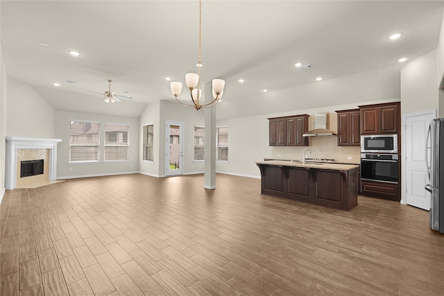 kitchen with lofted ceiling, a center island with sink, appliances with stainless steel finishes, hardwood / wood-style flooring, and wall chimney exhaust hood