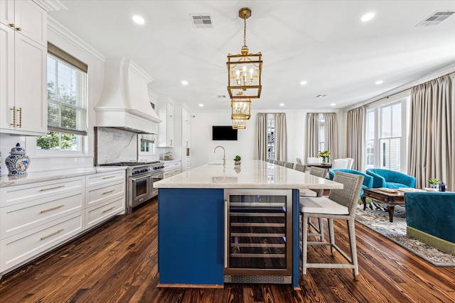 kitchen with white cabinets, beverage cooler, an island with sink, premium range hood, and range with two ovens