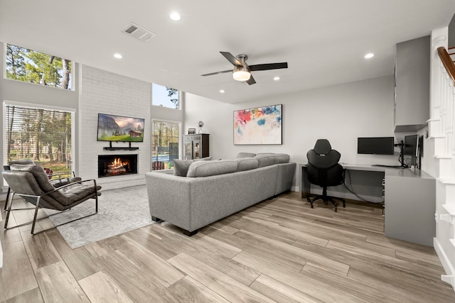 living room with built in desk, light hardwood / wood-style flooring, a brick fireplace, a towering ceiling, and ceiling fan