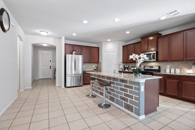 kitchen with an island with sink, stainless steel appliances, light stone counters, decorative backsplash, and light tile patterned floors