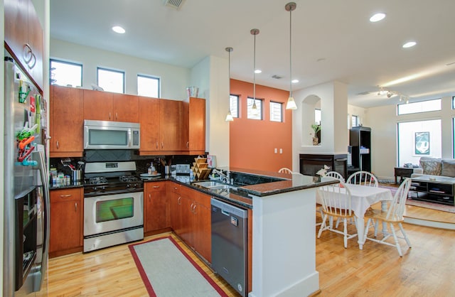 kitchen featuring appliances with stainless steel finishes, kitchen peninsula, hanging light fixtures, dark stone counters, and light hardwood / wood-style flooring