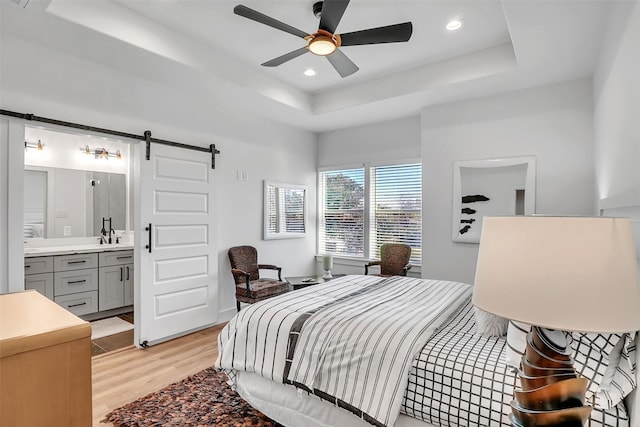 bedroom featuring ensuite bath, ceiling fan, a barn door, a tray ceiling, and light wood-type flooring