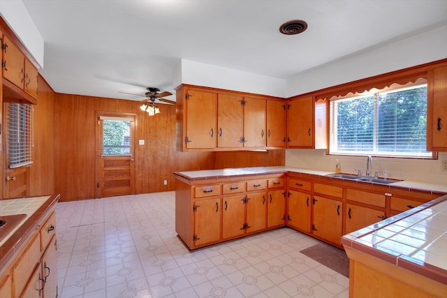 kitchen with kitchen peninsula, ceiling fan, tile countertops, wood walls, and sink