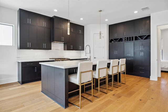 kitchen featuring light wood-type flooring, a kitchen bar, backsplash, hanging light fixtures, and a center island with sink