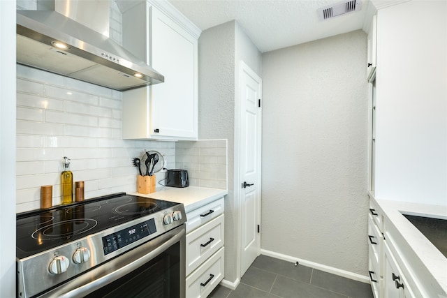 kitchen with stainless steel electric range oven, decorative backsplash, dark tile patterned flooring, wall chimney exhaust hood, and white cabinets