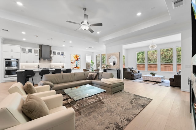 living room with a wealth of natural light, ceiling fan with notable chandelier, light wood-type flooring, and a raised ceiling