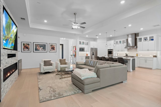 living room with ceiling fan, a stone fireplace, a tray ceiling, and light wood-type flooring