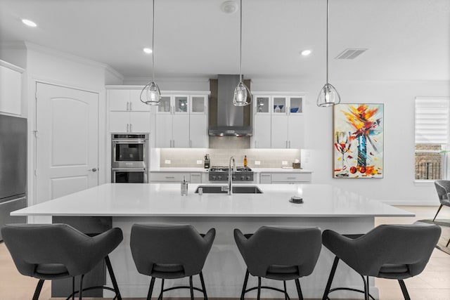 kitchen featuring wall chimney range hood, a center island with sink, sink, and appliances with stainless steel finishes