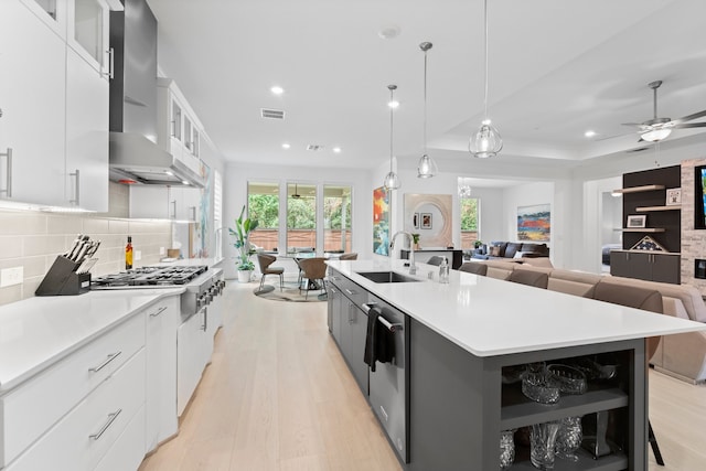 kitchen featuring wall chimney exhaust hood, an island with sink, backsplash, sink, and white cabinets