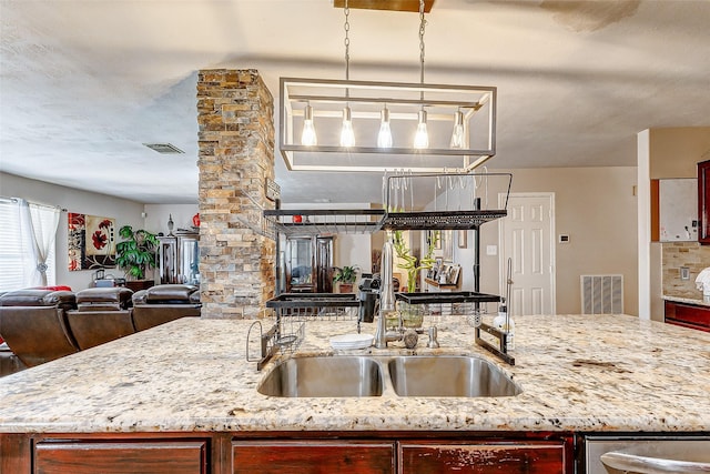 kitchen featuring sink, ornate columns, light stone counters, decorative light fixtures, and a center island with sink