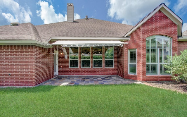 rear view of property featuring a chimney, a shingled roof, a patio area, a lawn, and brick siding