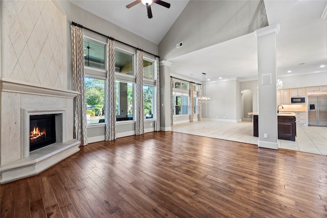 unfurnished living room with high vaulted ceiling, ceiling fan with notable chandelier, ornate columns, light hardwood / wood-style floors, and crown molding