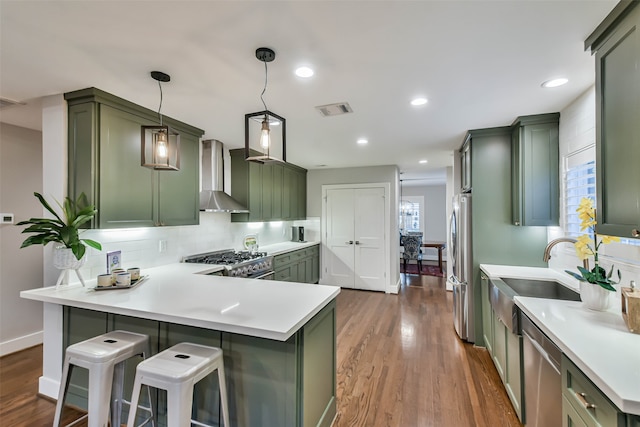 kitchen featuring appliances with stainless steel finishes, green cabinets, hanging light fixtures, a kitchen bar, and wall chimney exhaust hood