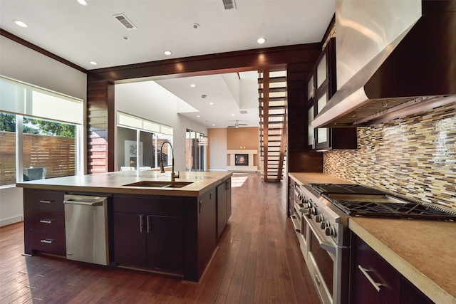 kitchen with wall chimney range hood, sink, high end stove, dark wood-type flooring, and a kitchen island with sink