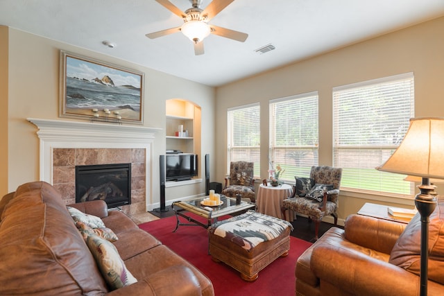 living room with a tile fireplace, built in features, and ceiling fan