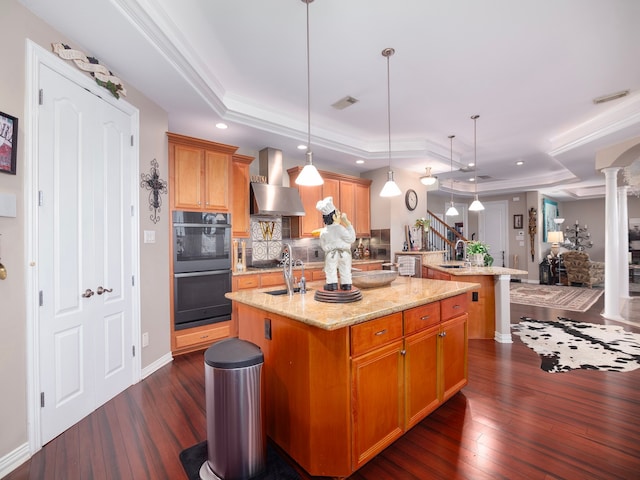 kitchen with ornate columns, wall chimney exhaust hood, a center island with sink, a raised ceiling, and dark hardwood / wood-style flooring