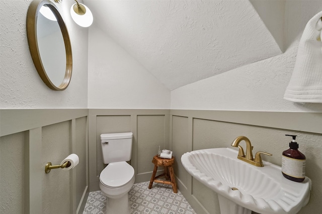 half bath with wainscoting, toilet, vaulted ceiling, a textured ceiling, and a sink