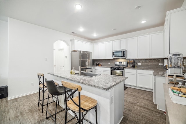 kitchen with light stone counters, stainless steel appliances, white cabinetry, an island with sink, and dark wood-type flooring