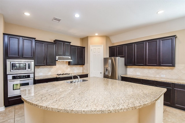 kitchen featuring sink, appliances with stainless steel finishes, tasteful backsplash, light stone counters, and a center island with sink