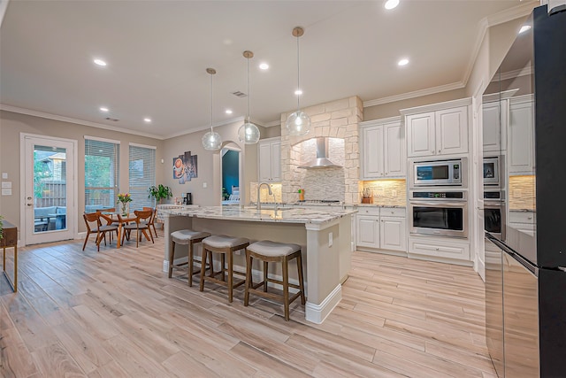 kitchen featuring wall chimney exhaust hood, white cabinetry, built in appliances, and an island with sink