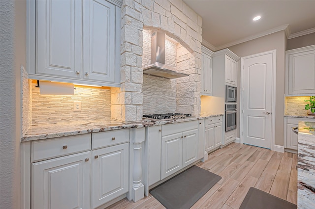 kitchen with wall chimney exhaust hood, ornamental molding, light stone countertops, white cabinets, and appliances with stainless steel finishes