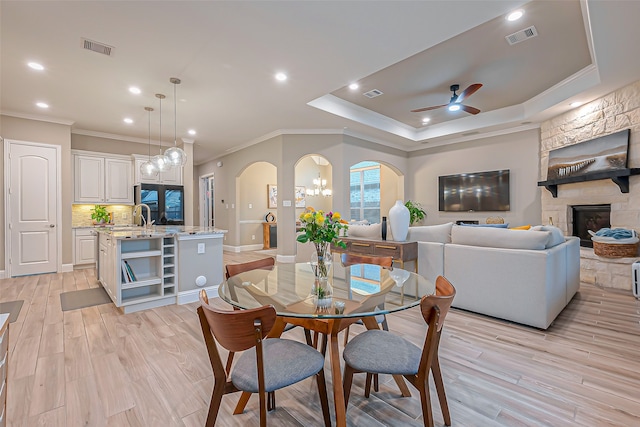 dining area with a stone fireplace, ornamental molding, light wood-type flooring, a raised ceiling, and ceiling fan