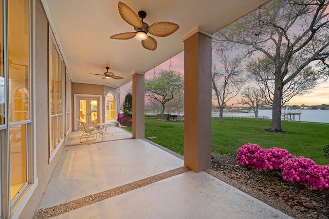 patio terrace at dusk featuring a water view, ceiling fan, a lawn, and french doors