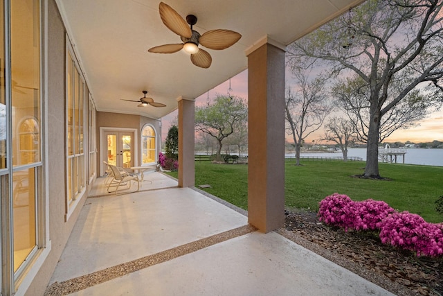 patio terrace at dusk featuring a lawn, a water view, ceiling fan, and french doors
