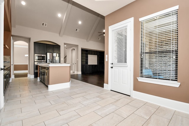 kitchen with an island with sink, appliances with stainless steel finishes, light stone countertops, light wood-type flooring, and lofted ceiling with beams