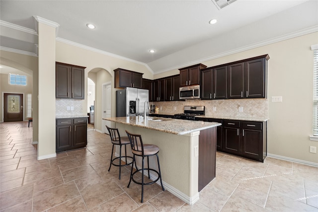 kitchen featuring a kitchen island with sink, backsplash, ornamental molding, dark brown cabinetry, and appliances with stainless steel finishes